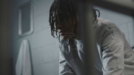 Foto de Primer plano de un afroamericano tratando de comer comida asquerosa de prisión sentado en la cama en la celda de la cárcel. Recluso adolescente o joven delincuente en un centro de detención juvenil o correccional. - Imagen libre de derechos