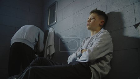 Téléchargez les photos : Un adolescent caucasien bouleversé est assis sur le lit en cellule de prison. Un détenu afro-américain se lave le visage dans l'évier. Les jeunes criminels purgent une peine d'emprisonnement en prison. Établissement correctionnel pour mineurs. - en image libre de droit
