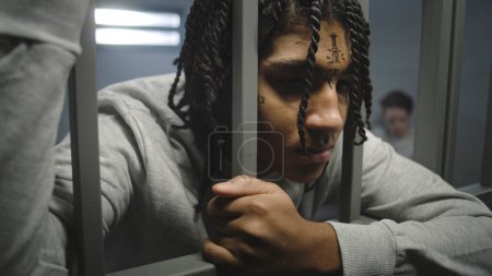 Foto de Primer plano de un prisionero adolescente afroamericano enojado con tatuajes en la cara parado en la celda de la prisión, sosteniendo barras de metal. Un joven recluso cumple condena de prisión por crimen en la cárcel. Centro de detención juvenil. - Imagen libre de derechos