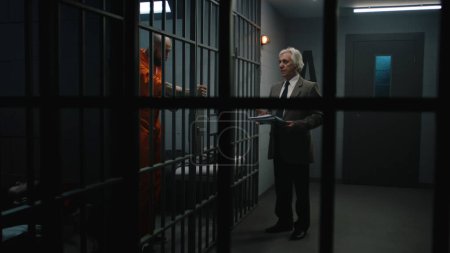 Criminal en uniforme naranja se apoya en las barras de celdas de la prisión, habla con el abogado y lee el contrato de abogado. El recluso cumple condena de prisión por delito en correccional. Gángster en centro de detención.