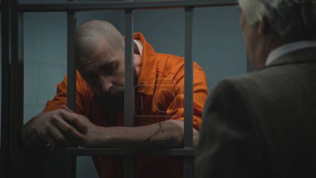 Prisoner in orange uniform leans on prison cell bars, talks with advocate and reads lawyer contract. Inmate serves imprisonment term for crime in correctional facility. Gangster in detention center.