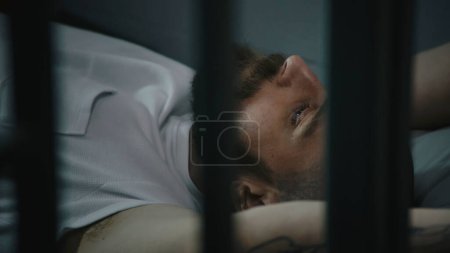 Photo for Criminal in orange uniform lies on prison cell bed. Prisoner serves imprisonment term for crime. Gangster in detention center, correctional facility. Justice system. View through metal bars. Close up. - Royalty Free Image
