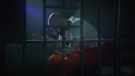 Criminal in orange uniform sits on bed in prison cell, stands up and looks at barred window. Prisoner serves imprisonment term for crime in jail. Gangster in detention center. View through metal bars.