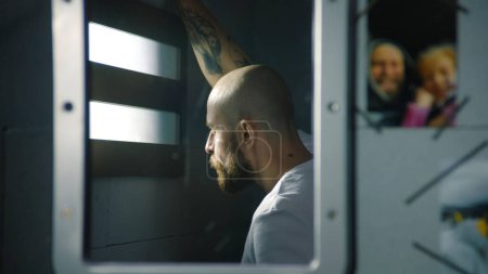 Reflection of male prisoner in mirror looking out the window in prison cell. Photos of family hang near the mirror. Inmate serves imprisonment term in jail. Detention center or correctional facility.