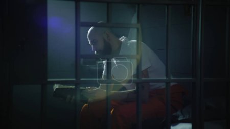 Téléchargez les photos : Un prisonnier en uniforme orange est assis sur son lit dans une cellule de prison, lit la Bible. Un homme condamné illégalement purge une peine d'emprisonnement. Vue à travers des barres métalliques. Centre de détention ou établissement correctionnel. - en image libre de droit