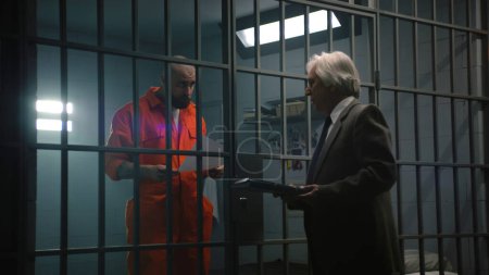 Foto de Criminal en uniforme naranja está detrás de las rejas en la celda de la prisión, habla con el abogado y lee contrato. El preso cumple condena de prisión por delito en correccional. Gángster en centro de detención. - Imagen libre de derechos
