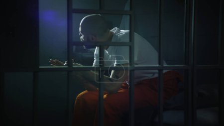 Photo for Criminal in orange uniform sits on bed in prison cell, stands up and looks at barred window. Prisoner serves imprisonment term for crime in jail. Gangster in detention center. View through metal bars. - Royalty Free Image