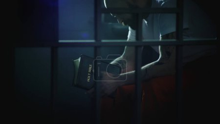 Photo for Male prisoner in orange uniform sits on bed in prison cell, reads Bible. Illegally convicted man serves imprisonment term in jail. View through metal bars. Detention center or correctional facility. - Royalty Free Image