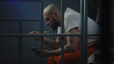 Male prisoner in orange uniform sits on bed in prison cell, eats disgusting prison food from iron bowl. Inmate serves imprisonment term for crime in jail. Detention center or correctional facility.