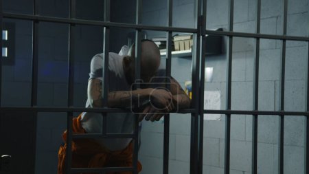 Photo for Depressed male prisoner in orange uniform stands in prison cell leaning and holding hands on metal bars. Inmate serves imprisonment term for crime in jail. Detention center or correctional facility. - Royalty Free Image