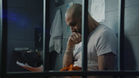 Un prisionero deprimido de uniforme naranja mira los dibujos de su hijo y llora sentado en la cama en la celda de la prisión. Criminal cumple pena de prisión en la cárcel. Centro de detención o correccional.