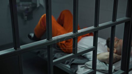 Photo for Inmate in orange uniform lies on prison cell bed. Prisoner serves imprisonment term for crime. Criminal in detention center, correctional facility. Justice system. View through metal bars. Dolly shot. - Royalty Free Image