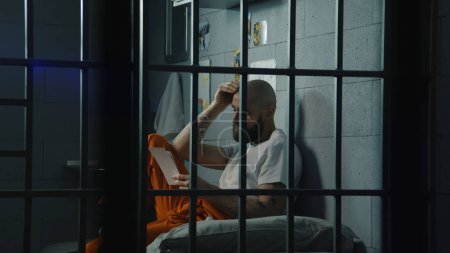 Un prisionero de uniforme trastornado ve fotos de familia y niños sentados en la cama en la celda de la prisión. Un hombre condenado ilegalmente cumple condena de prisión en prisión. Centro de detención o correccional.