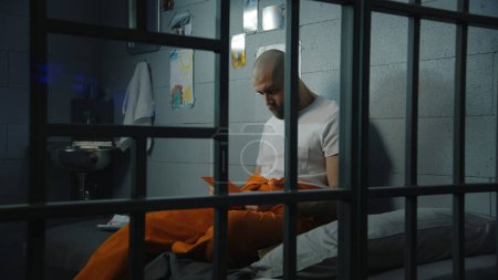 Foto de Hombre prisionero en uniforme naranja se sienta en la cama en la celda de la prisión, mira las fotografías de la familia y los niños. Criminal cumple pena de prisión por crimen en la cárcel. Centro de detención o correccional. - Imagen libre de derechos