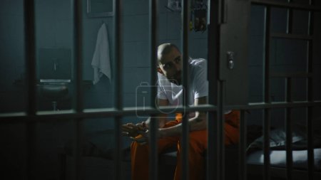 Foto de Criminal en uniforme naranja se sienta en la cama de la celda de la prisión. El preso cumple condena de prisión por crimen en la cárcel. Gángster en el centro de detención, correccional. Sistema de justicia. Vista a través de barras de metal. - Imagen libre de derechos