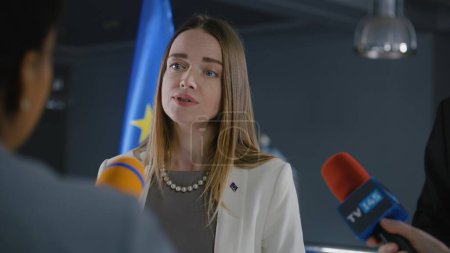 Positive female Representative of the European Union talks to news journalists, gives interview for media and television in government building. Inspirational speech of politician at press conference.