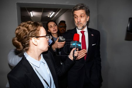 Photo for Busy politician answers press questions, gives interview for news in corridor of government building. Confident diplomat surrounded by crowd of journalists. Political speech during press conference. - Royalty Free Image