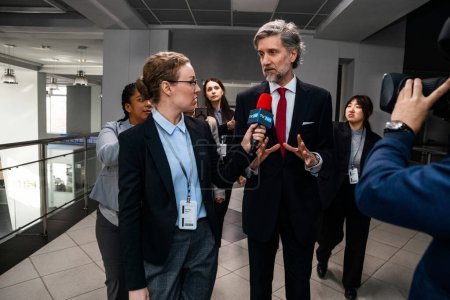 Busy politician answers press questions, gives interview for news in corridor of government building. Confident diplomat surrounded by crowd of journalists. Political speech during press conference.