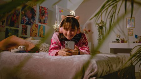 Téléchargez les photos : Asiatique adolescente dans un casque parle par chat vidéo, surfe sur Internet en utilisant un téléphone mobile tout en étant allongé sur le lit dans une chambre confortable. Fille passer du temps libre et s'amuser à la maison. Concept de style de vie. - en image libre de droit
