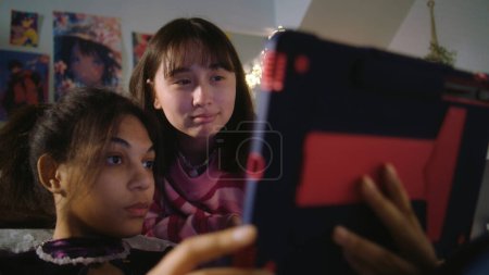Téléchargez les photos : Asiatique adolescent mensonges sur lit et montres content avec ami. Afro-Américaine s'assoit au sol, surfe sur Internet à l'aide d'une tablette. Les filles multiculturelles passent du temps libre ensemble à la maison. Relation amicale. - en image libre de droit