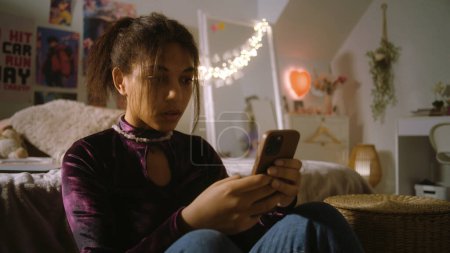 Photo for Teenager sits on floor near bed in her room screams and emotionally types on phone. Angry African American girl communicates using phone. Spending leisure time at home. Home with stylish interior. - Royalty Free Image