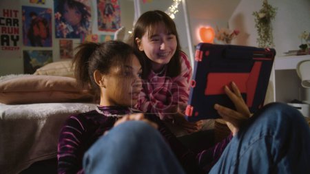 Afroamerikanerin sitzt am Boden, surft mit Tablet im Internet. Mongolischer Teenager liegt im Bett, schaut zufrieden mit Freund zu. Multi-ethnische Mädchen verbringen gemeinsam ihre Freizeit zu Hause. Beziehung zu Freunden.