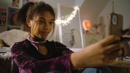 Photo for Beautiful teenager sits on floor near bed in her room and takes a pictures using phone. African American girl chats with friends or creates blog content. Spending leisure time at comfortable home. - Royalty Free Image