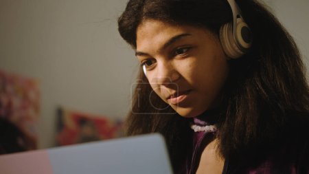 Photo for African American girl in headphones sits in her room and surfs internet using laptop. Beautiful teenager chats with friends, watches video content. Spending leisure time at comfortable home. Close up. - Royalty Free Image