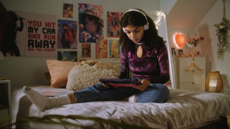 Photo for African American girl in headphones sits on bed in her room and surfs internet using laptop. Beautiful teenager chats with friends or watches video content. Spending leisure time at comfortable home. - Royalty Free Image