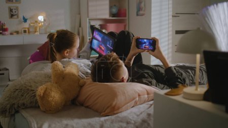 Foto de Dos adolescentes yacen en la cama en su habitación y juegan videojuegos en dispositivos digitales. Chica utiliza la tableta, niño utiliza el teléfono para jugar juntos. Hermana pasar tiempo libre con su hermano. Relación familiar. - Imagen libre de derechos