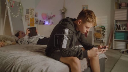 Photo for Young girl lies on bed in room and scrolls social networks using tablet. Boy with phone in hands plays games. Teenagers spending leisure time. Family relationship. Home with cozy and stylish interior. - Royalty Free Image