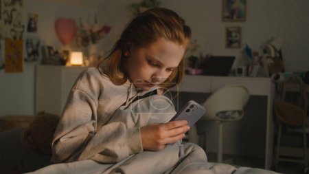Photo for Caucasian teenager gets up early in morning in her comfort bedroom. Young girl takes her mobile phone and turns off alarm. Teen sits on cozy bed and uses her smartphone to chat in social network. - Royalty Free Image