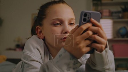 Photo for Close up of Caucasian teenager using her mobile phone to chat with friends in social networks and surfs the Internet. Young girl lies on her cozy bed in light bedroom. Teen spends weekend at home. - Royalty Free Image