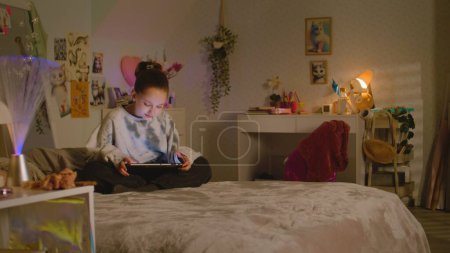 Photo for Young girl sits on bed in her room and surfs the internet or chats online using tablet. Cute schoolgirl spending leisure time at home scrolling social networks. Home with cozy and stylish interior. - Royalty Free Image