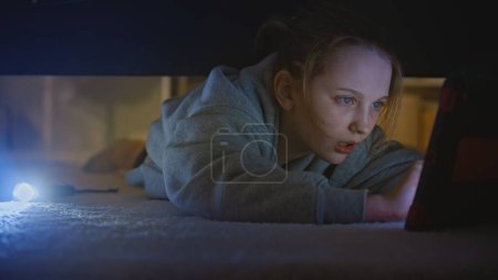 Kaukasischer Teenager kommt in modernen hellen Raum. Junge schöne Mädchen mit Taschenlampe versucht, digitale Tablet-Computer unter großem Bett in ihrem Schlafzimmer zu finden. Schießen unter dem Bett. Lifestyle-Konzept.