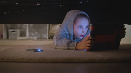 Junges Mädchen kommt mit Taschenlampe auf ihrem Handy ins große helle Schlafzimmer. Kaukasischer Teenager legt sich unter das Bett, findet einen digitalen Tablet-Computer und nutzt ihn. Schießen unter dem Bett. Lifestyle-Konzept.