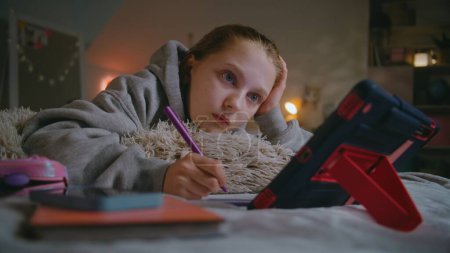 Photo for Young girl lies on bed in comfort bedroom, writes in notebook and does school lessons using tablet computer. Caucasian teenager spends time and studies online at home in daytime. Lifestyle concept. - Royalty Free Image