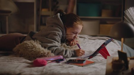 Photo for Young girl lies on bed at home, writes in notebook and does online homework for school using tablet computer. Caucasian teenager spends time in cozy and comfort bedroom in daytime. Lifestyle concept. - Royalty Free Image