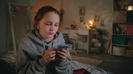 Photo for Young beautiful girl uses smartphone, sets up sound in headphones, then talks with friend sitting on bed in cozy bedroom. Caucasian teenager spends leisure time at home in daytime. Lifestyle concept. - Royalty Free Image