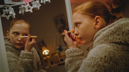 Foto de Joven hermosa chica hace maquillaje de las cejas de pie delante del espejo en el acogedor y confortable dormitorio. Adolescente caucásica pasar el tiempo libre y divertirse en casa por la noche. Concepto de estilo de vida - Imagen libre de derechos