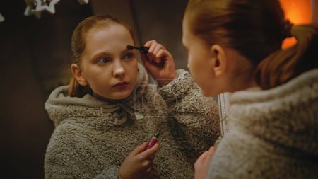 Photo for Young Caucasian girl does eyebrows makeup standing and looking in the mirror in cozy and comfort bedroom. Teenage girl spending leisure time and having fun at home at night. Concept of lifestyle. - Royalty Free Image