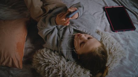Photo for Young girl in headphones lies on the bed at home, listens to music and surfs the Internet using mobile phone. Caucasian teenager spends leisure time in comfort bedroom in daytime. Lifestyle concept. - Royalty Free Image