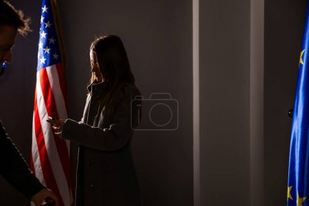 Téléchargez les photos : La silhouette d'une femme politique avec un smartphone entre les mains se dresse entre les drapeaux des États-Unis et de l'UE - en image libre de droit