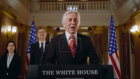 Photo for Positive President of the United States gives inspirational speech in the White House, then leaves. Senior US minister speaks at press campaign for media and television. American flags in background. - Royalty Free Image