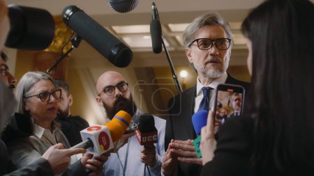 Politician answers press questions and gives interview for television news. Mature diplomat surrounded by crowd of journalists in parliament hall. Press conference. Inspirational speech. Slow motion.