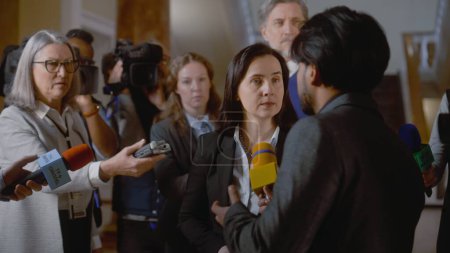 Politician answers press questions and gives interview on television political program. Female diplomat surrounded by crowd of journalists. Political speech during press conference. Hot button issues.
