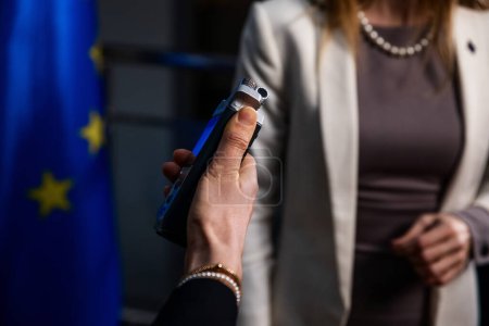 Téléchargez les photos : Vue rapprochée d'un enregistreur de voix dans la main d'un journaliste destiné à une femme politique arborant le drapeau de l'UE - en image libre de droit