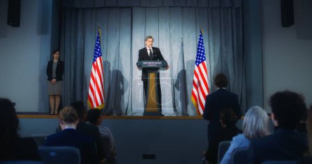 Confident American republican politician delivers successful speech to supporters at government election rally. New president of USA gives interview for media. Backdrop with American flags. Zoom out.