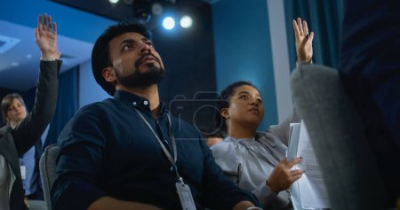 Photo for Multiethnic press workers sit in the conference room. Indian male journalist raises hand, asks question and writes down the answer during press campaign with organization representative or politician. - Royalty Free Image