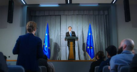 Photo for Confident politician makes an announcement, answers media questions and gives interview. Mature representative of the European Union during performance at press conference. Backdrop with EU flags. - Royalty Free Image